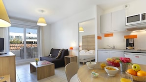 A modern hotel room with a sofa, a dining table with breakfast items, a kitchen with white cabinets, and a balcony with a view of the city. - Vacancéole - Cap Camargue (Le Grau-Du-Roi)