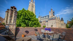 A rooftop terrace with a view of a historic building, furnished with wicker chairs and a table with flowers.