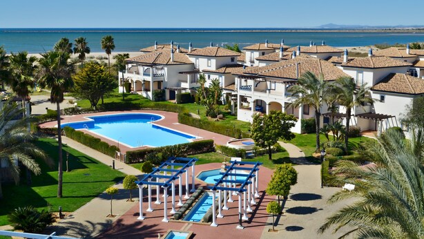 A resort with a large swimming pool, palm trees, and a view of the sea. - Leo Apartamentos San Bruno (Ayamonte)