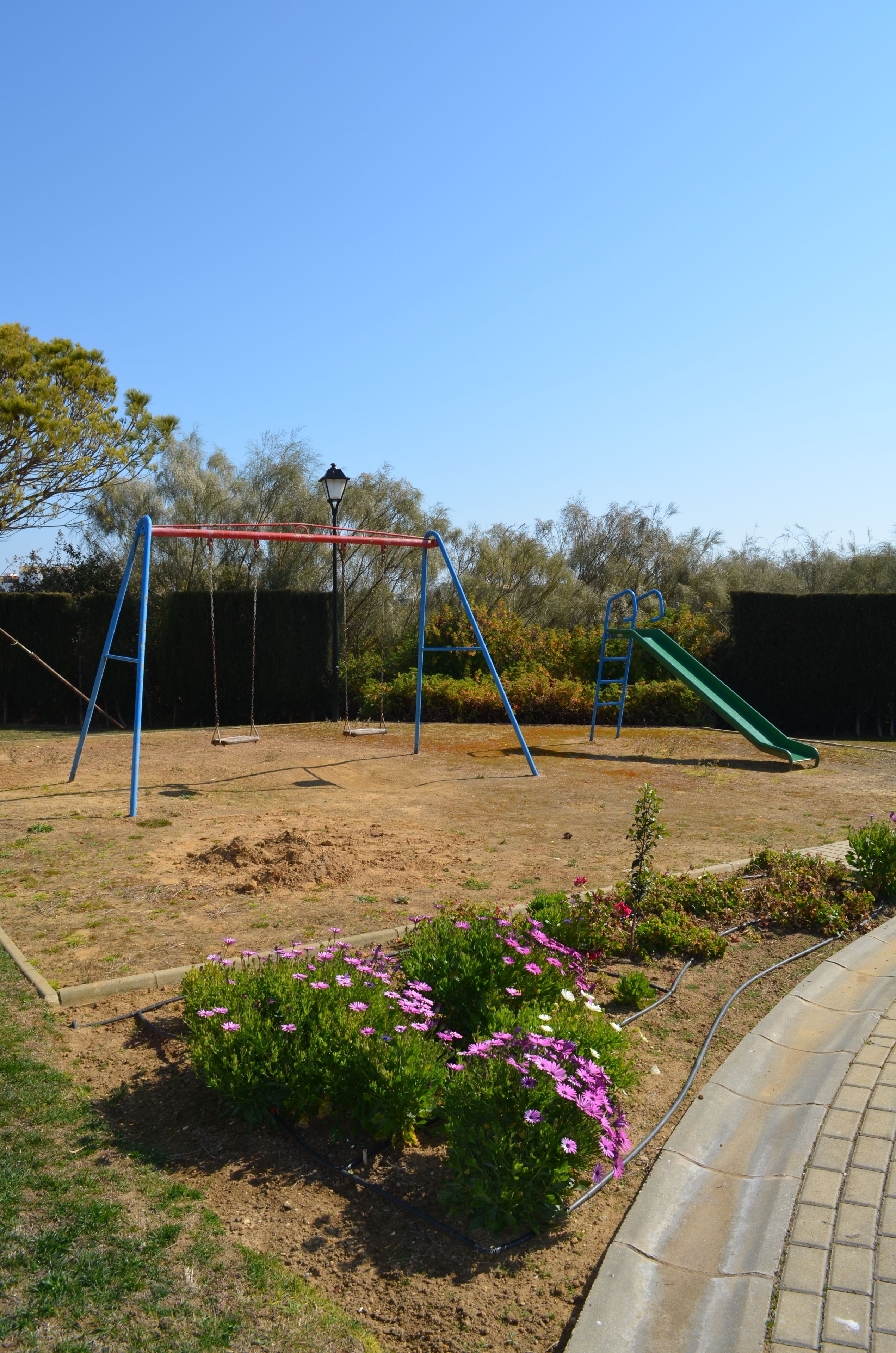 Un parque infantil con columpios y un tobogán, rodeado de un jardín con flores moradas.