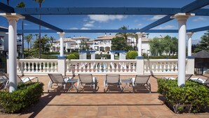 A covered outdoor seating area with white columns and a tiled floor, overlooking a pool and buildings. - Leo Apartamentos San Bruno (Ayamonte)