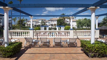 Un espacio de estar cubierto al aire libre con columnas blancas y piso de baldosas, con vista a una piscina y edificios.