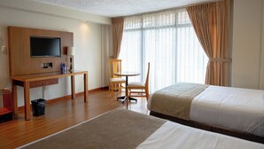 Una habitación de hotel con dos camas, un escritorio con televisor, una silla y una ventana con cortinas.