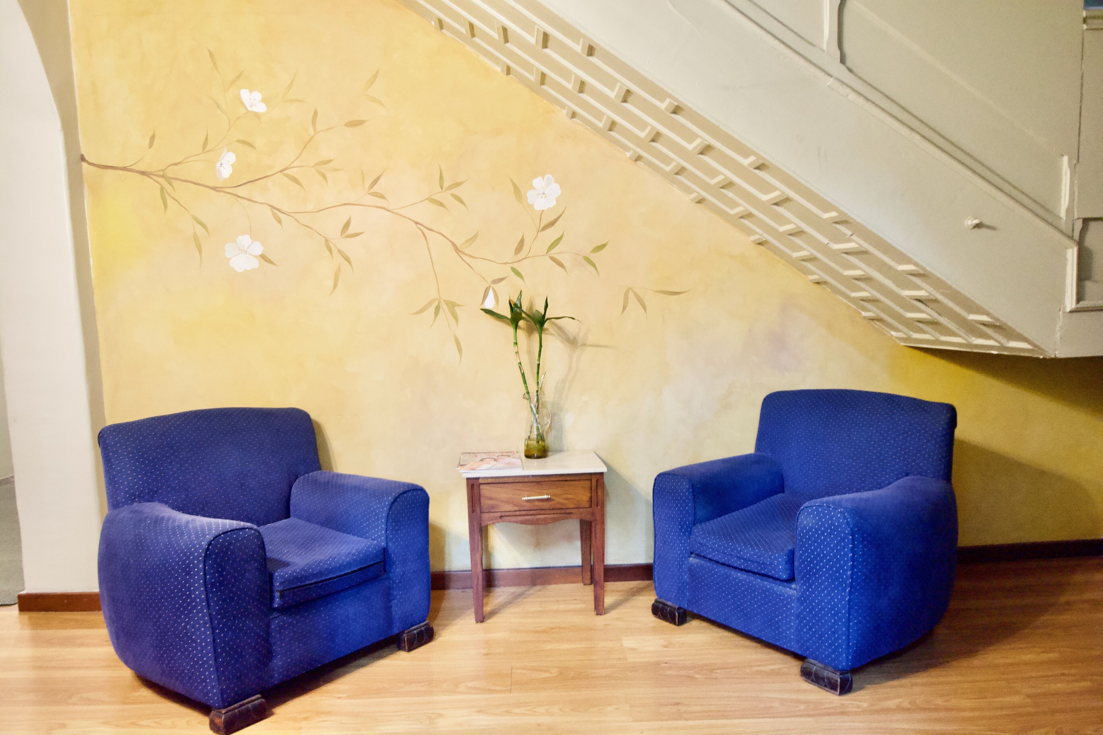 Een kamer met twee blauwe fauteuils, een klein houten bijzettafeltje met een vaas, en een muur met een muurschildering van takken en bloemen.