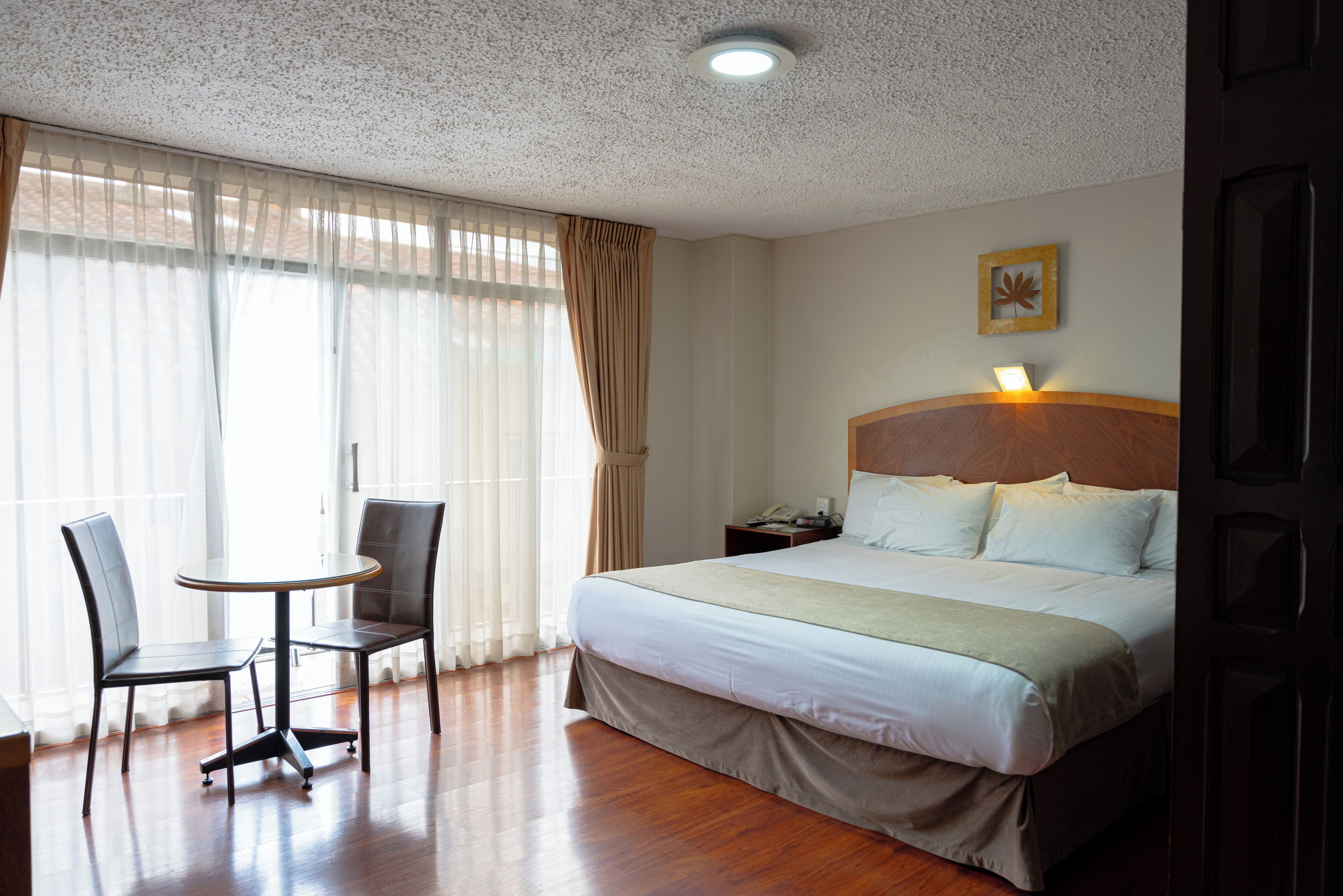 A hotel room with a bed, two chairs, a small table, a nightstand, and a wall-mounted lamp.