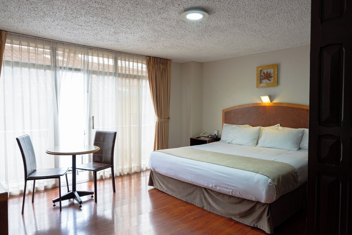 Una habitación de hotel con cama, dos sillones, una mesita, una mesita de noche y una lámpara de pared.