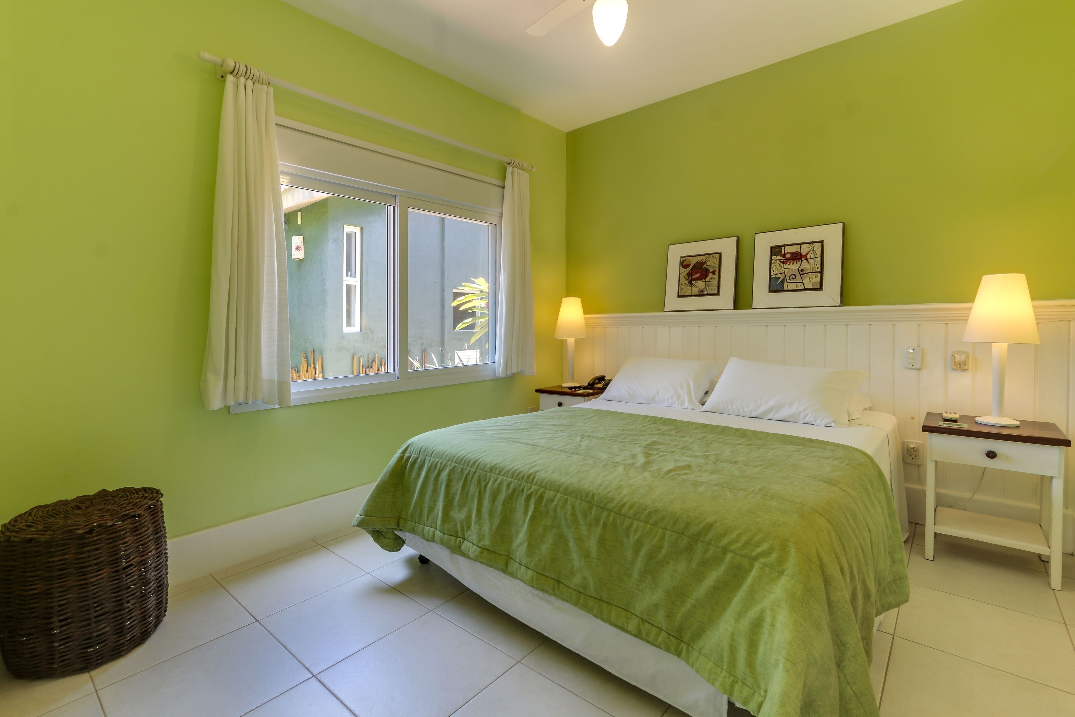 Un dormitorio con cama, dos mesitas de noche con lámparas, una ventana con cortinas y una pared decorada con cuadros.
