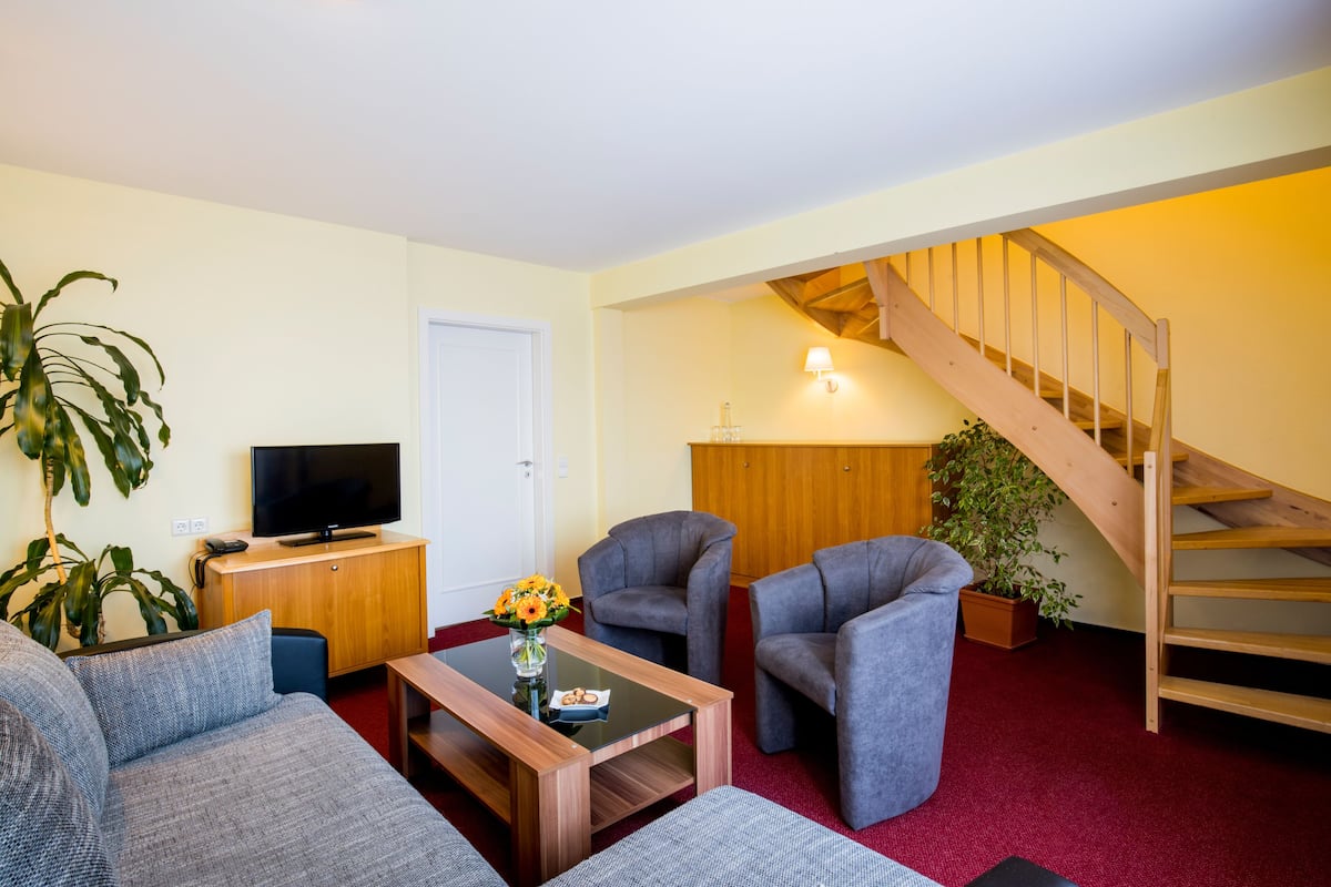 A living room with a sofa, two armchairs, a coffee table with a vase of flowers, a television on a wooden cabinet, and a staircase.