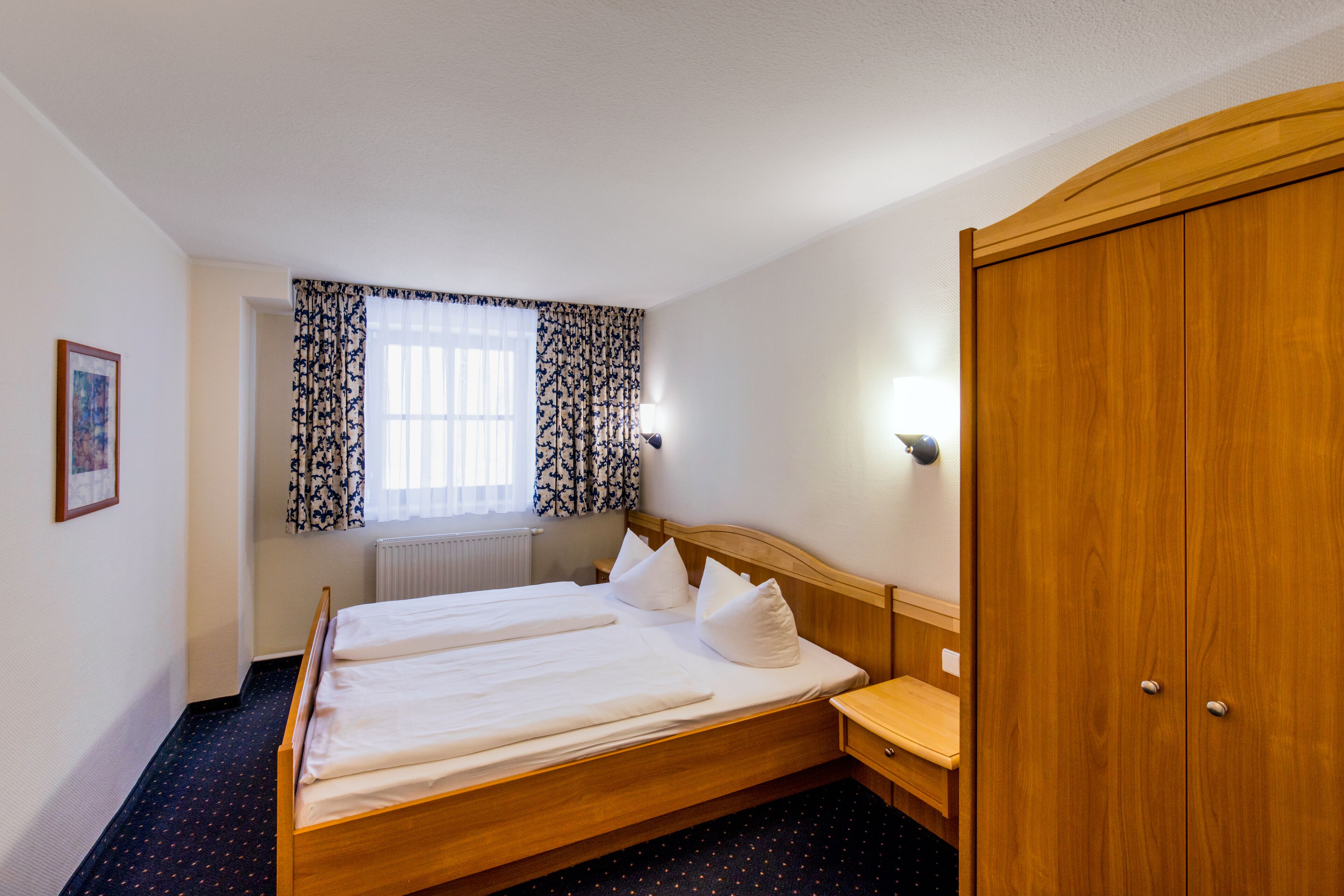 A hotel room with a double bed, a wooden wardrobe, a bedside table, a window with patterned curtains, and a radiator.