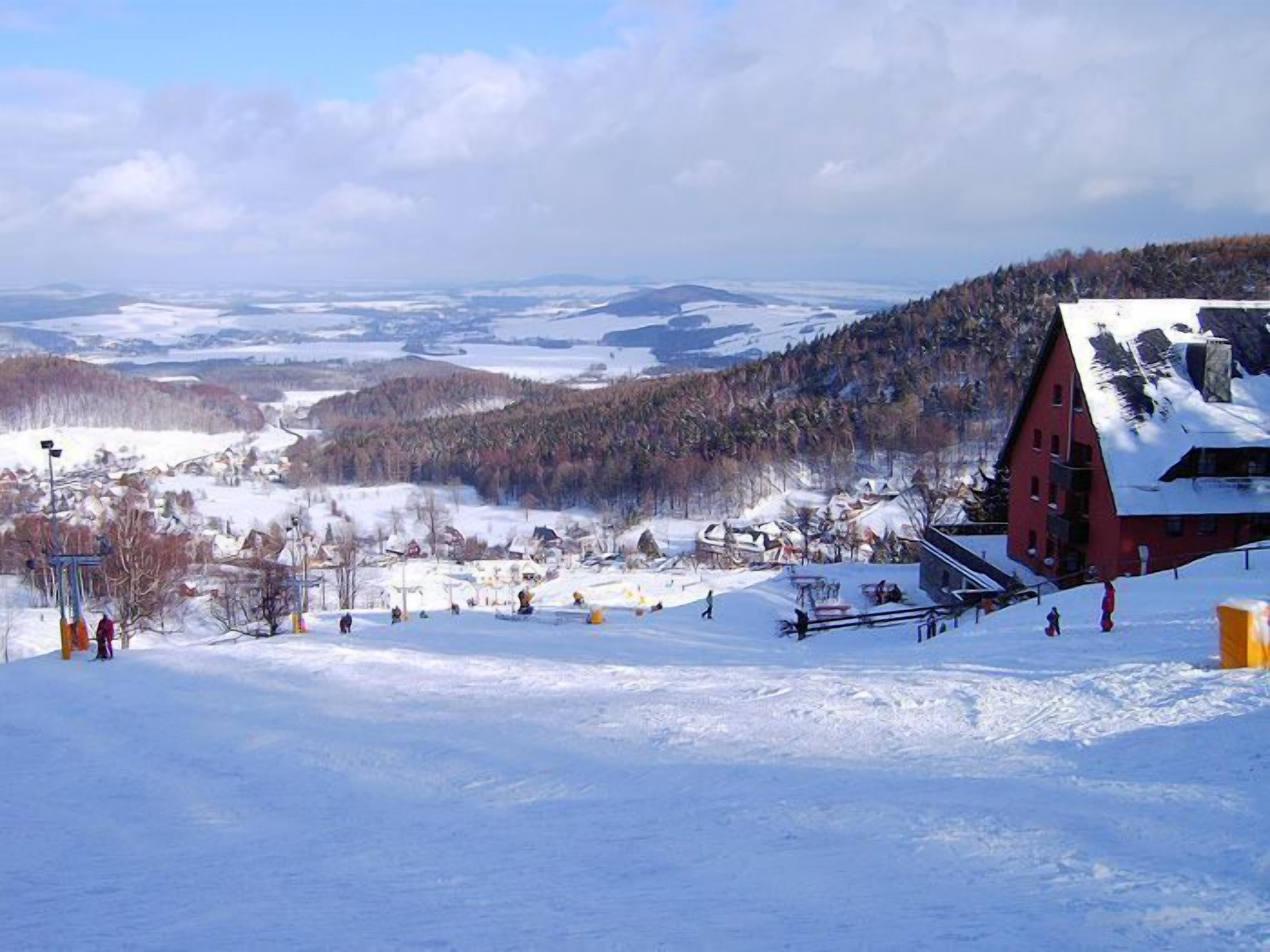 A snow-covered ski resort with a red building, ski slopes, and a mountainous backdrop.