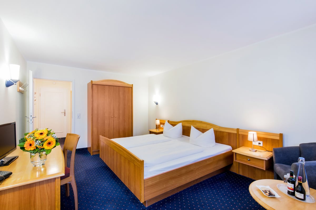 A hotel room with a wooden bed, a desk with a vase of flowers, a chair, a TV, and a nightstand with a lamp.