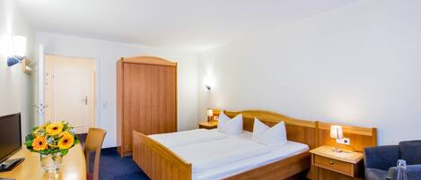 Une chambre d’hôtel avec un lit en bois, un bureau agrémenté d’un vase de fleurs, une chaise, une télévision et une table de chevet avec une lampe.