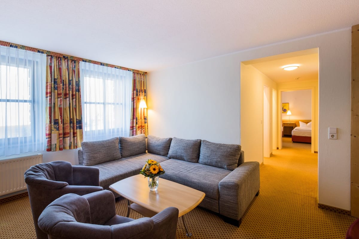 A hotel room with a grey sofa, two armchairs, a small wooden coffee table with a vase of flowers, a bed with a white duvet, and a nightstand with a lamp.