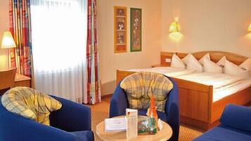 A hotel room with a bed, two blue armchairs, a small table with a bottle and a menu, and a window with curtains.