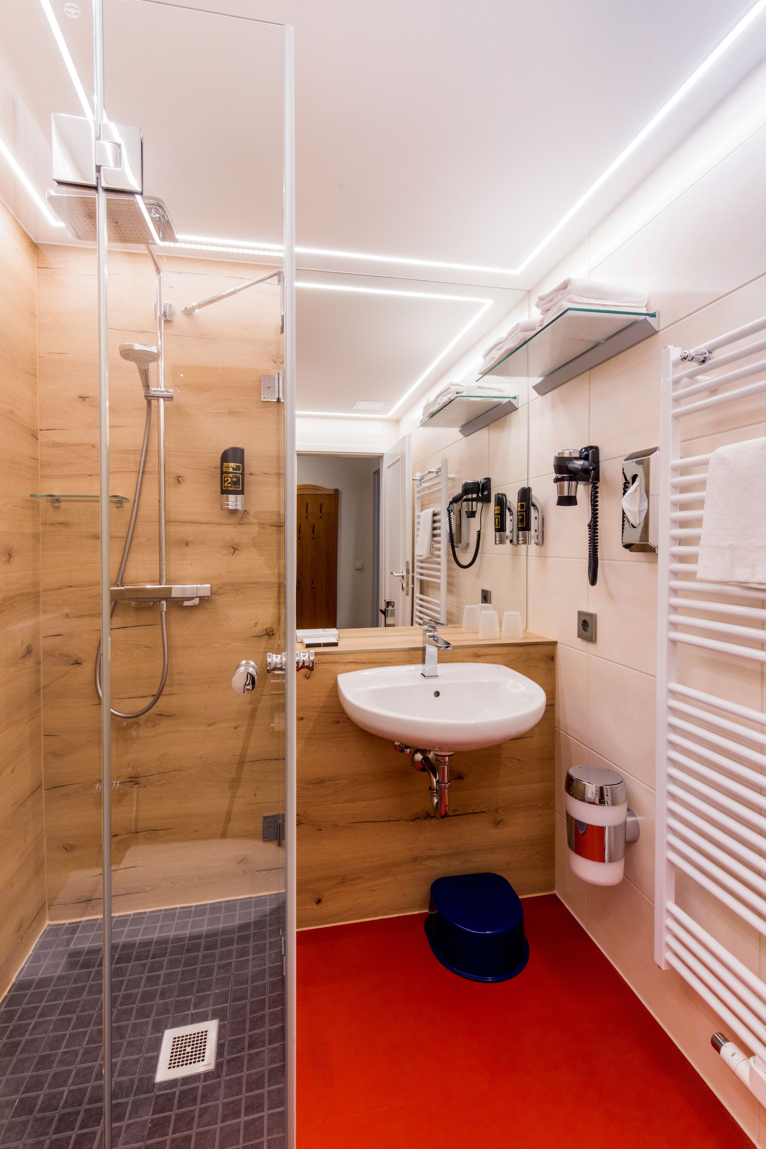 A modern bathroom with a glass shower enclosure, a wooden vanity, a sink, and a red floor.