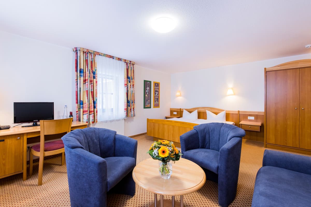 A hotel room with a wooden desk, two blue armchairs, a round table with a vase of flowers, a bed with wooden headboard, and a wardrobe.