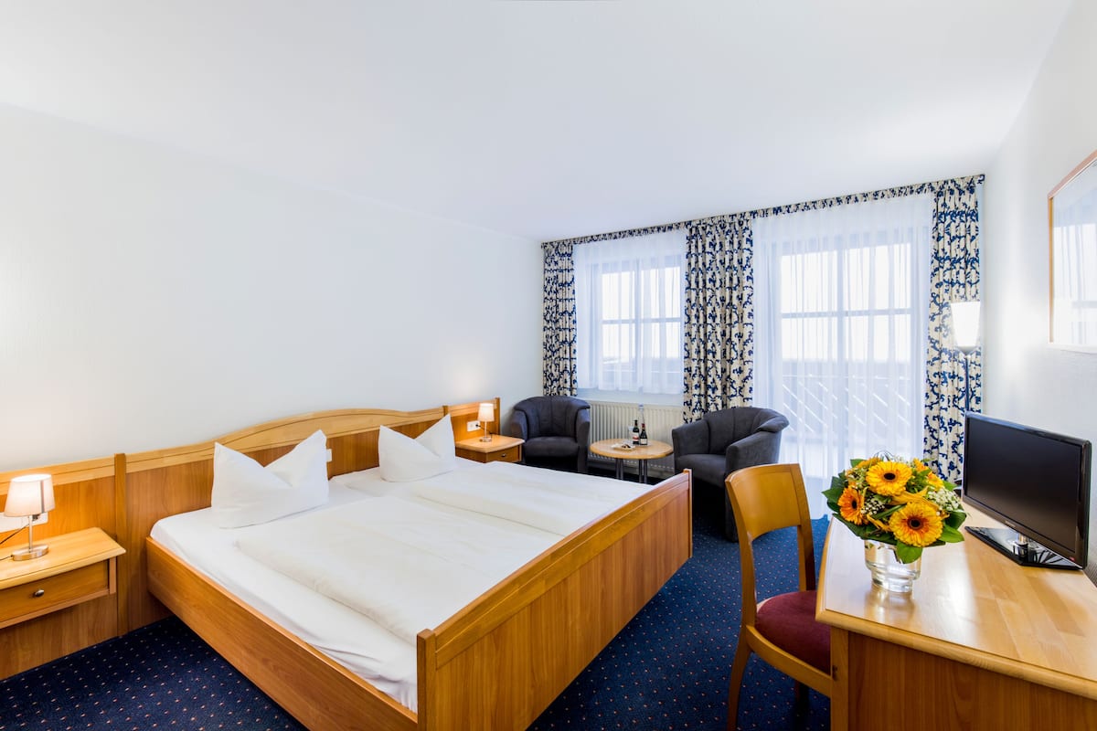 A hotel room with a wooden bed, a desk with a computer, two armchairs, a small table with a lamp, and a vase with flowers.