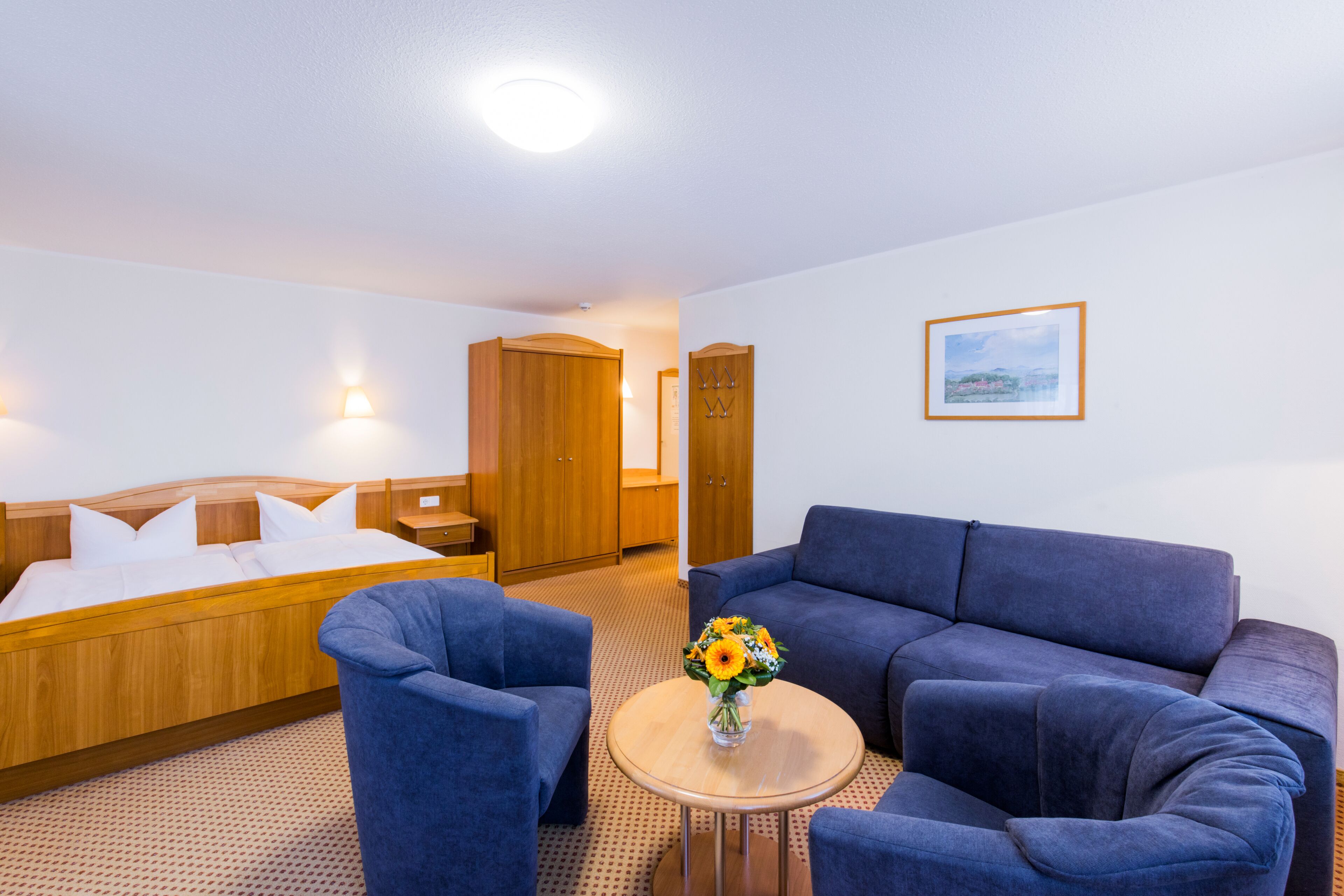 A hotel room with a bed, two blue armchairs, a small round table with a vase of flowers, a wooden wardrobe, and a framed picture on the wall.
