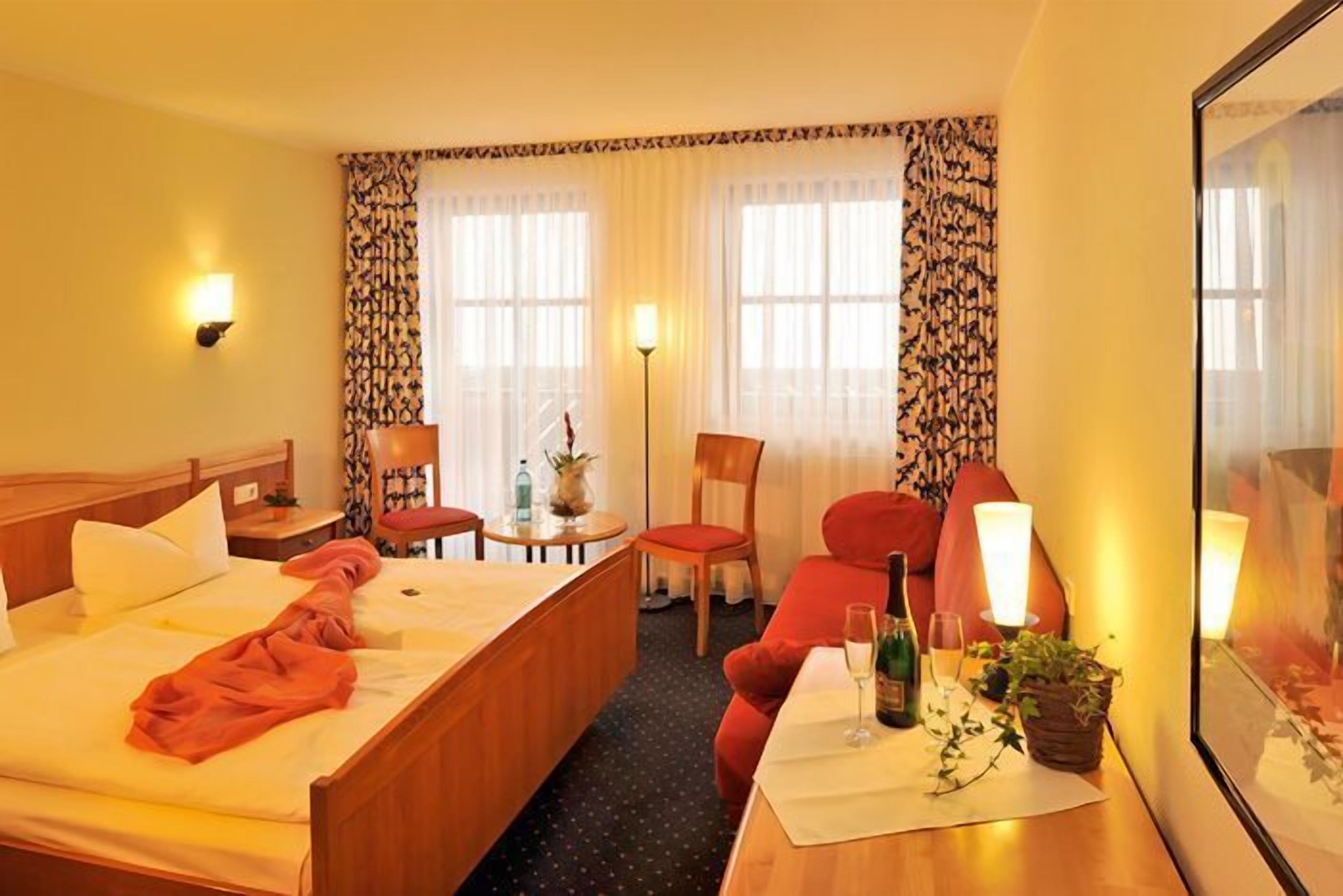 A hotel room with a bed, a desk with a bottle and glass, a chair, a small table, and a lamp.