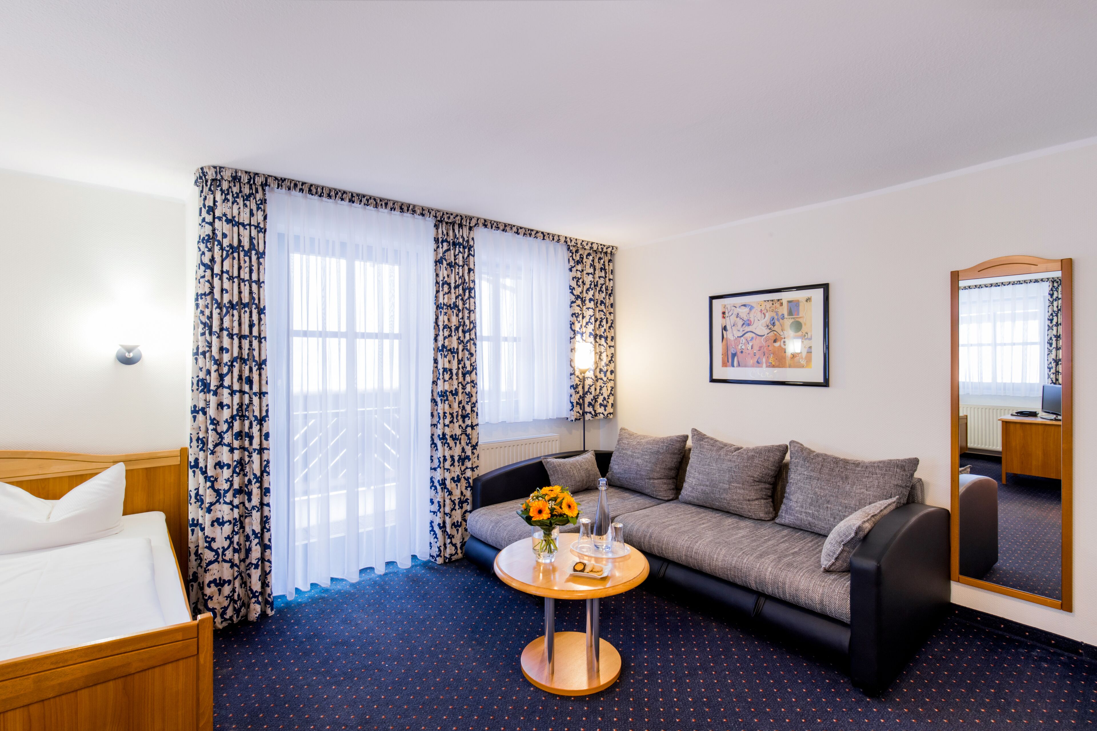 Una habitación de hotel con una cama, un sofá, una mesa redonda con un florero y un espejo grande.