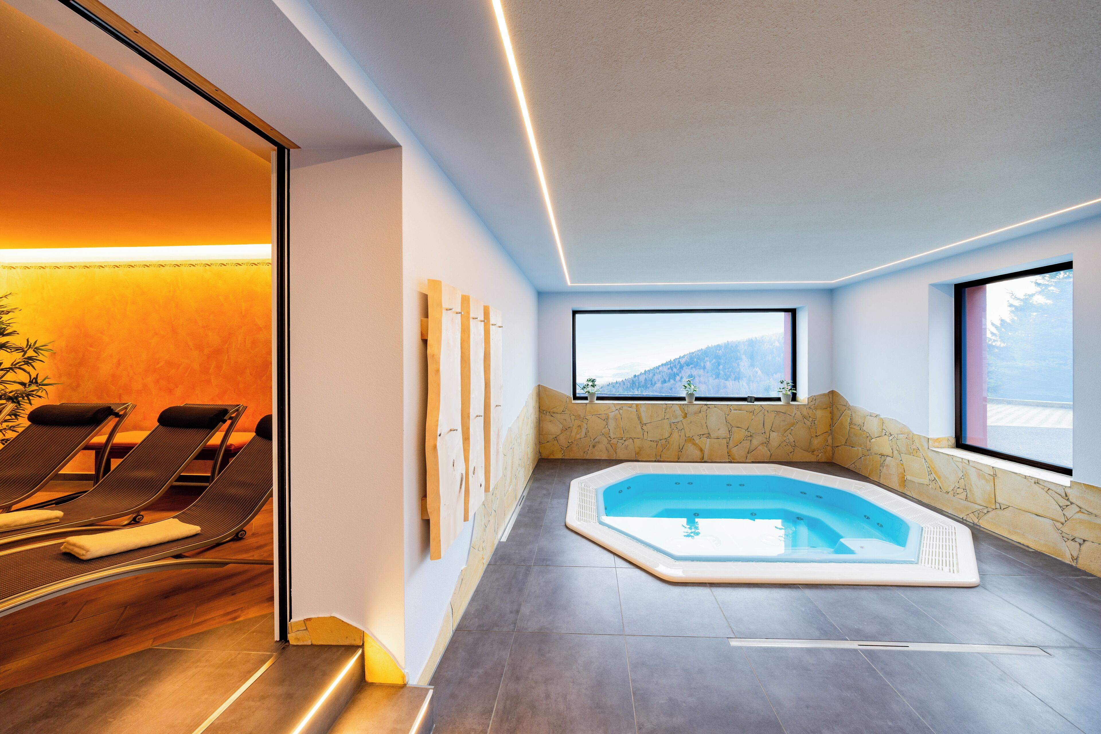 Ein moderner Innenpoolbereich mit Whirlpool, Liegestühlen und Blick auf einen Berg durch große Fenster.