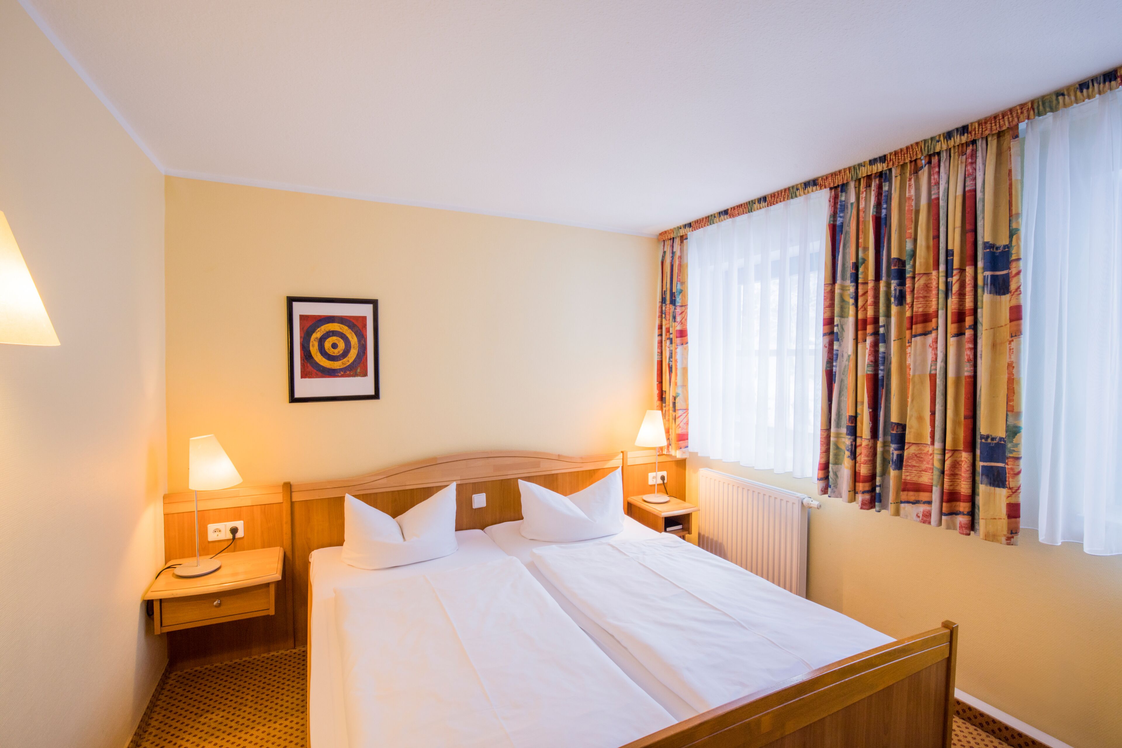 A hotel room with a double bed, two bedside tables with lamps, a framed picture on the wall, and a window with colorful curtains.
