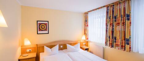 A hotel room with a double bed, two bedside tables with lamps, a framed picture on the wall, and a window with colorful curtains.
