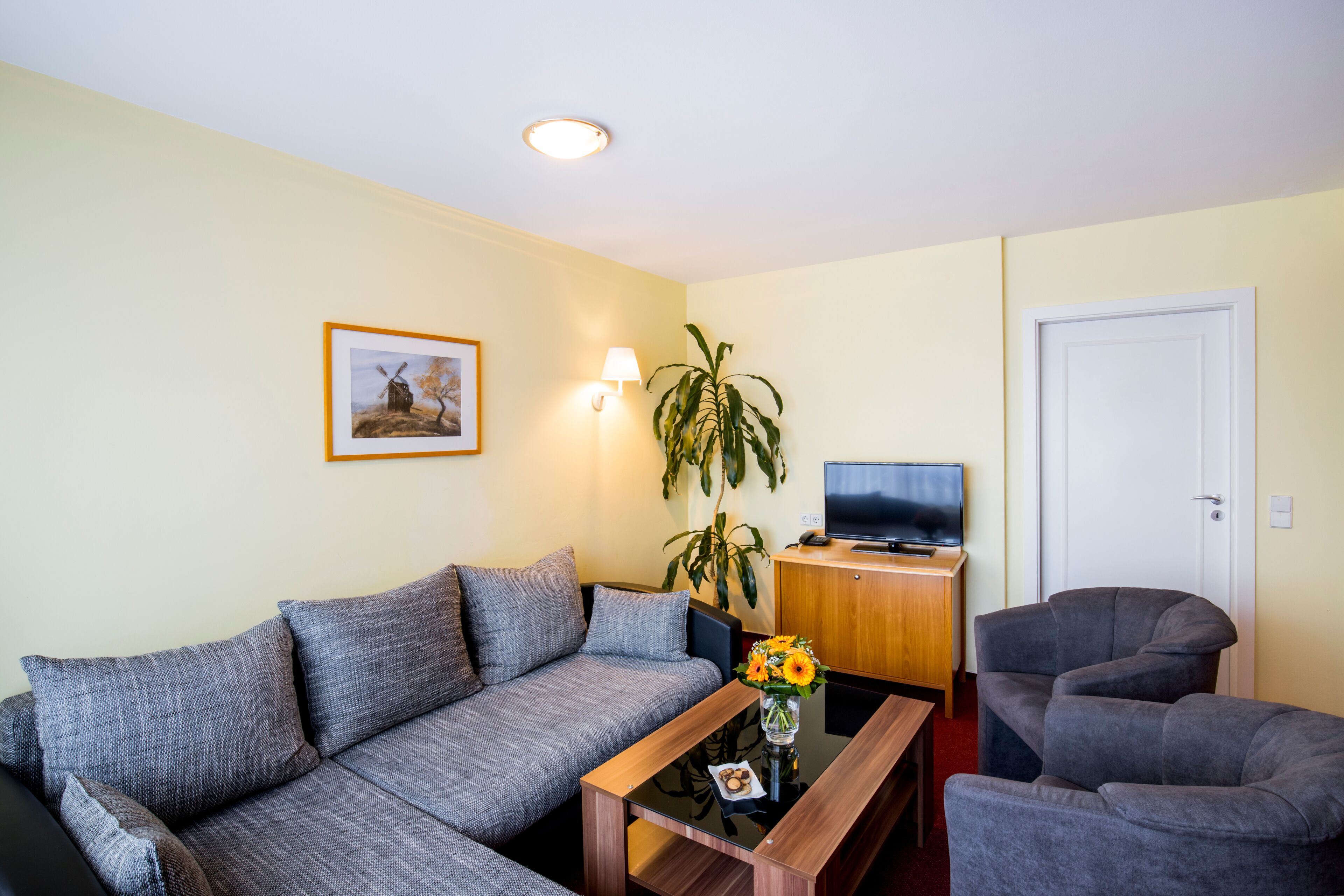 Una sala de estar con un sofá gris, una mesa de centro con un jarrón de flores, un televisor sobre un mueble de madera y una puerta.