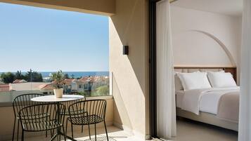 Un balcón con mesa y sillas, una cama con ropa blanca y vista al mar y edificios.