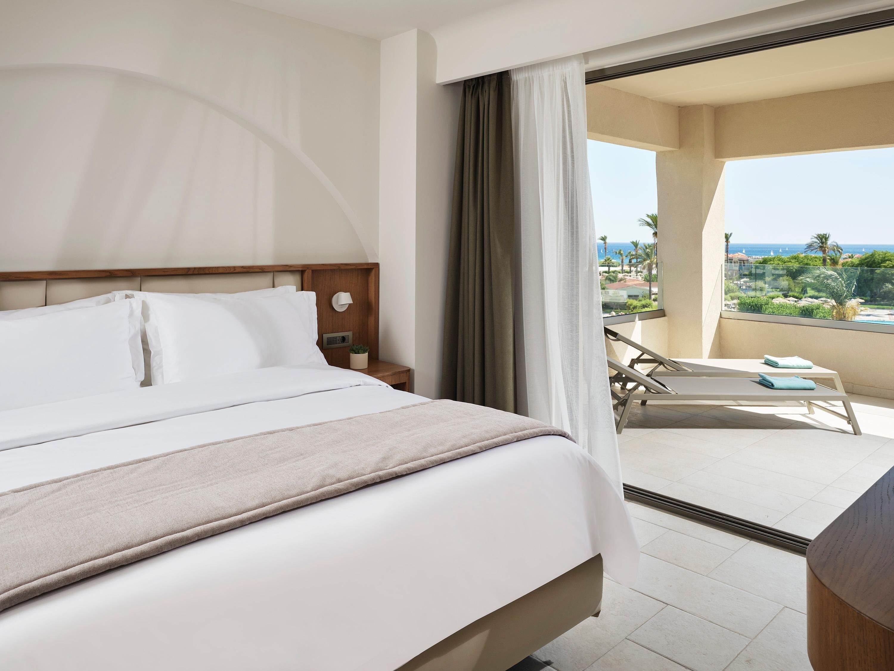 A hotel room with a bed, a nightstand, a lamp, and a view of a balcony with a pool and palm trees.