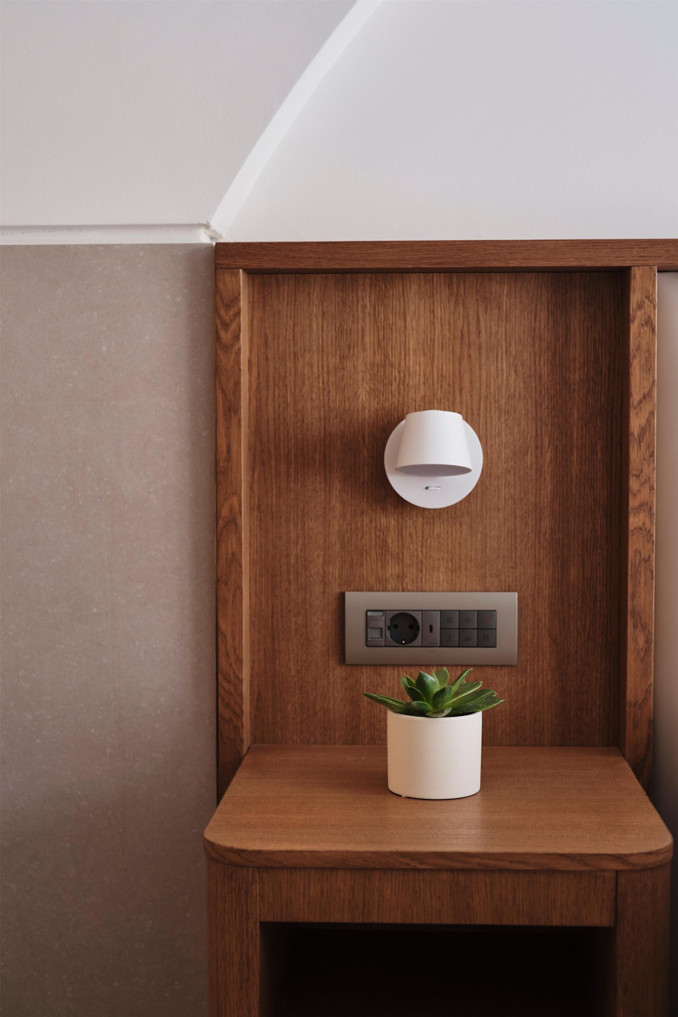 A corner with a wooden shelf, a potted plant, a wall-mounted light, and a control panel.