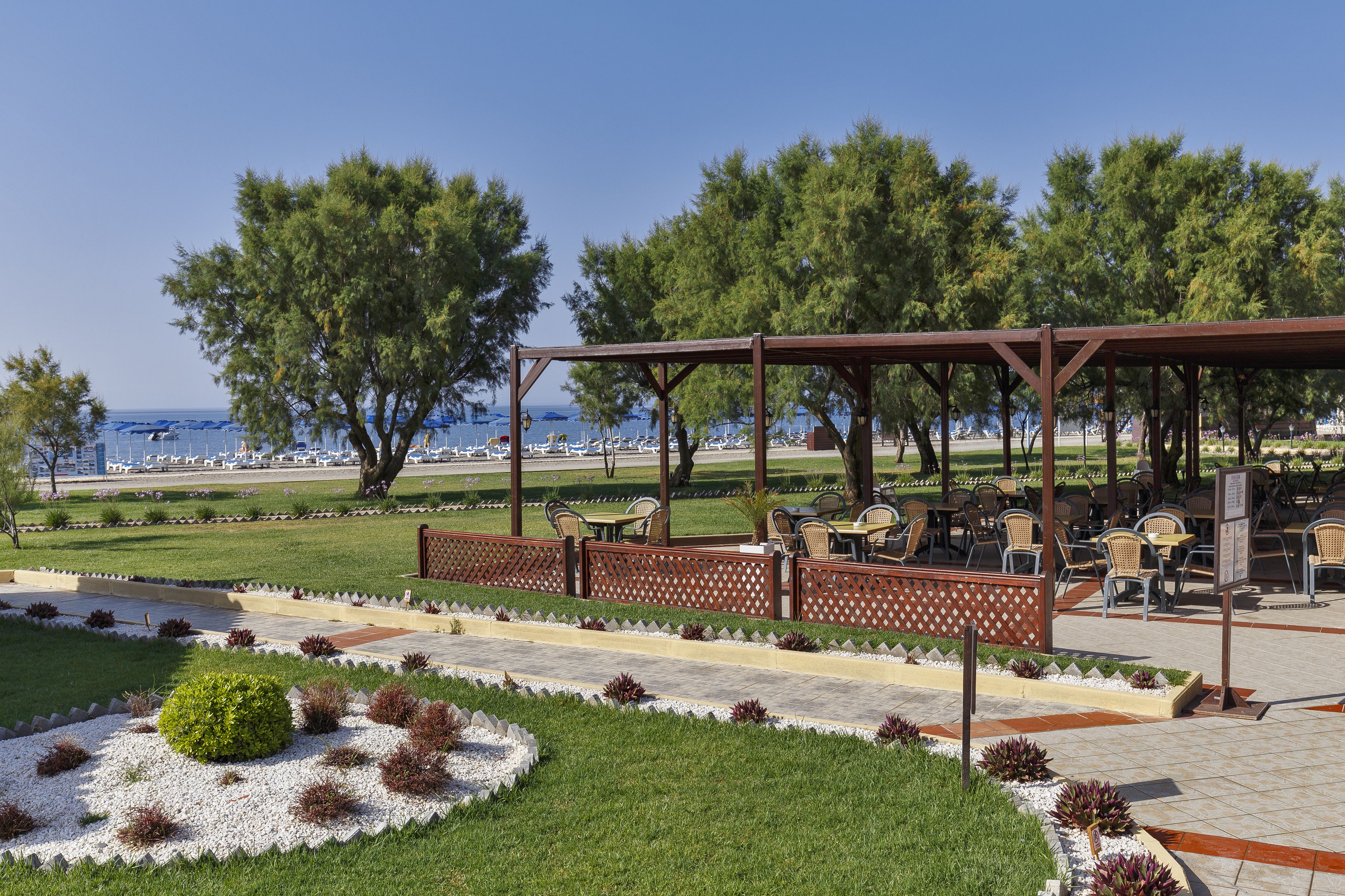 Ein überdachter Außenbereich mit Tischen und Stühlen, ein angelegter Garten mit einem Busch im Zentrum sowie Meer- und Bootsblick im Hintergrund.