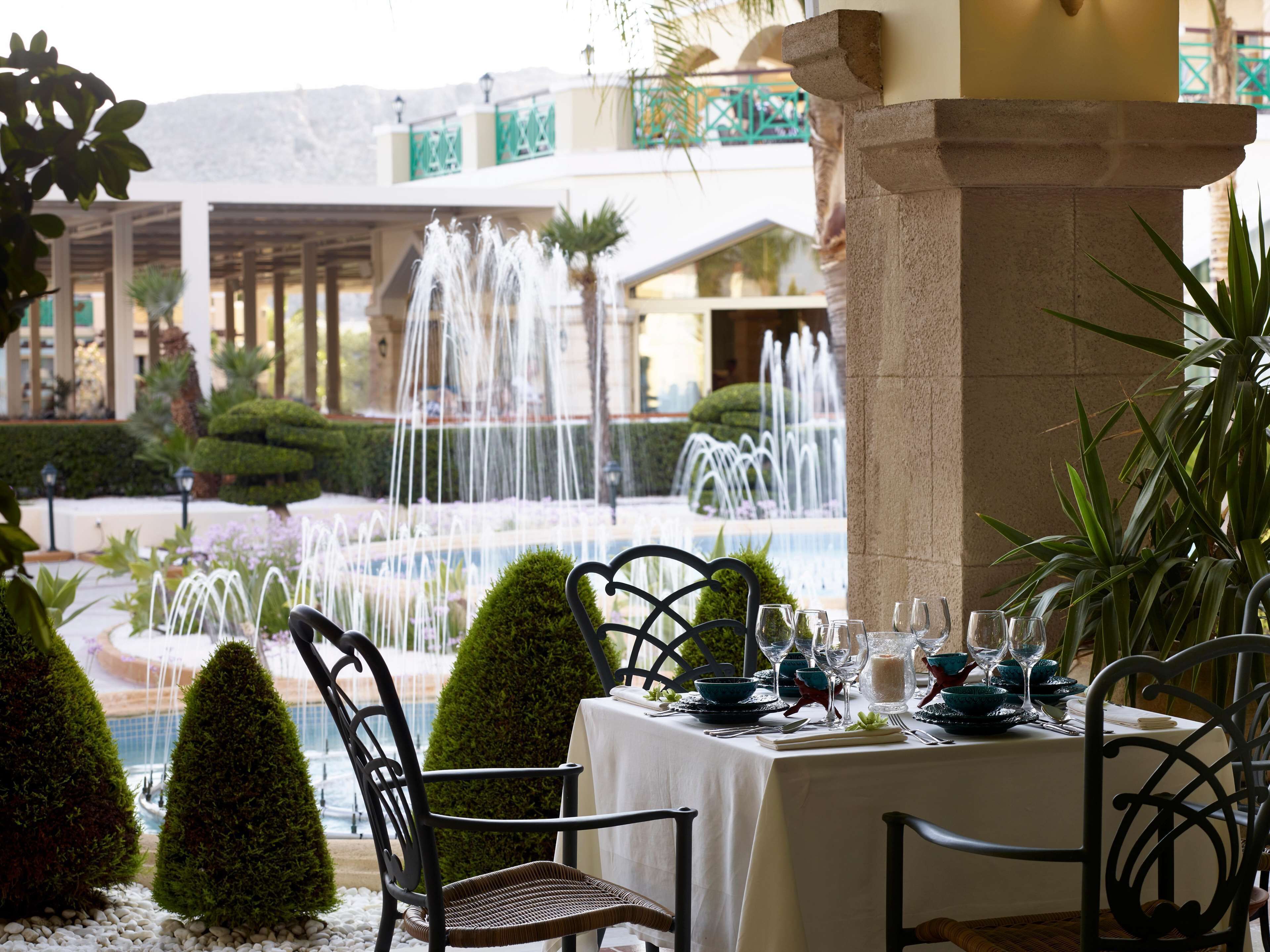 A dining table set with chairs, a fountain in the background, and a building with balconies.