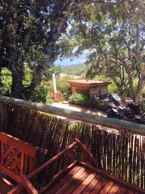 A wooden deck with a bamboo fence, overlooking a small wooden building and a scooter, with trees and a distant view.