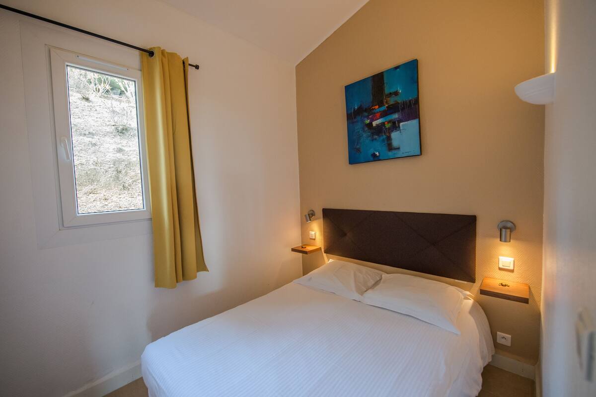 Ein ordentlich gemachtes Bett mit weißer Bettwäsche, ein Fenster mit gelben Vorhängen, ein Bild an der Wand und Nachttischlampen.