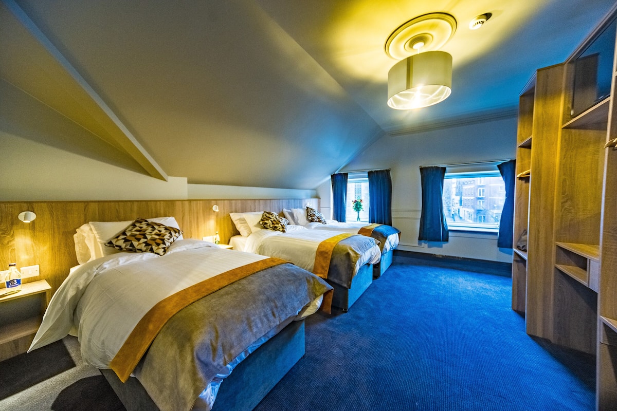 A hotel room with three beds, a large window with curtains, a wooden wardrobe, and a carpeted floor.