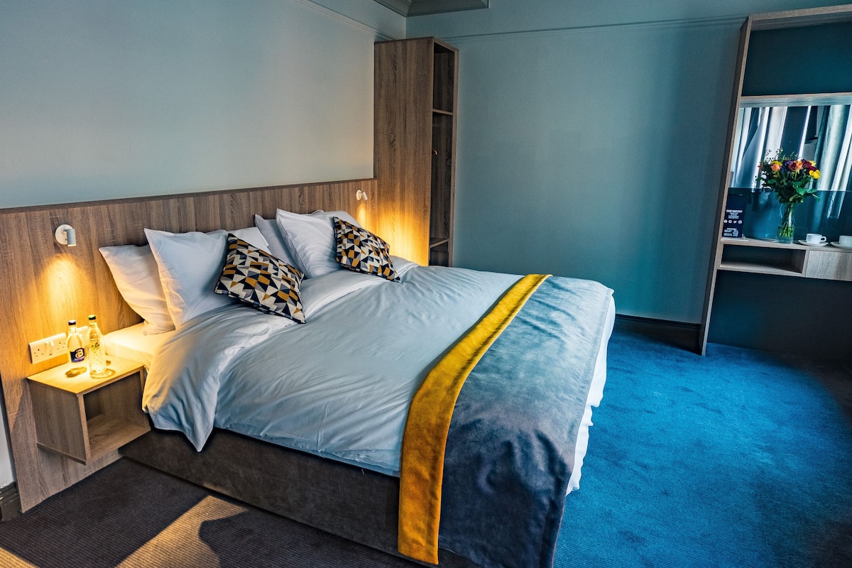 A neatly made bed with a blue and yellow bedspread, a wooden headboard, and a nightstand with a lamp and bottles.