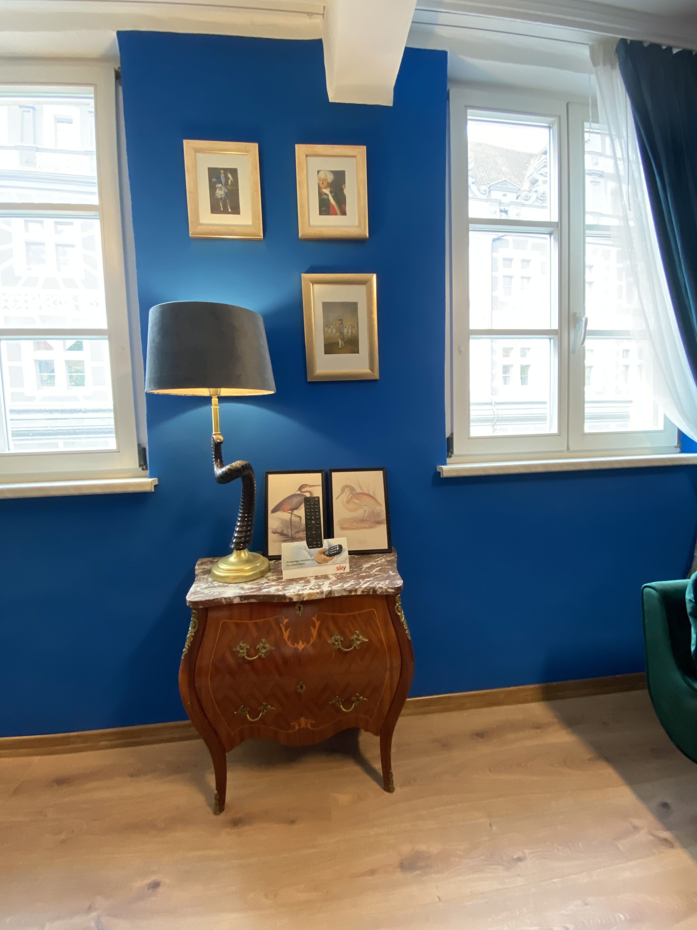 A room with a blue wall, a wooden cabinet with a lamp, framed pictures, and a window with curtains.