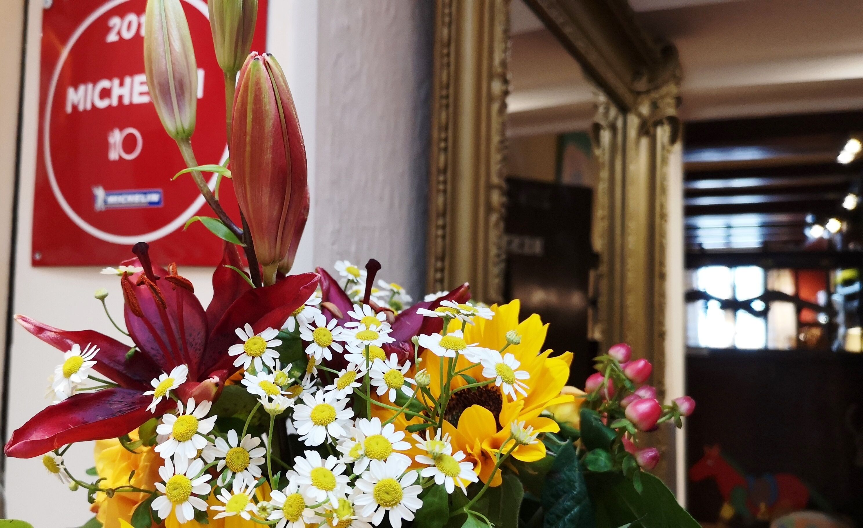 Ein Blumenstrauß mit roten, gelben und weißen Blüten, im Hintergrund ein rotes Schild mit weißer Schrift.