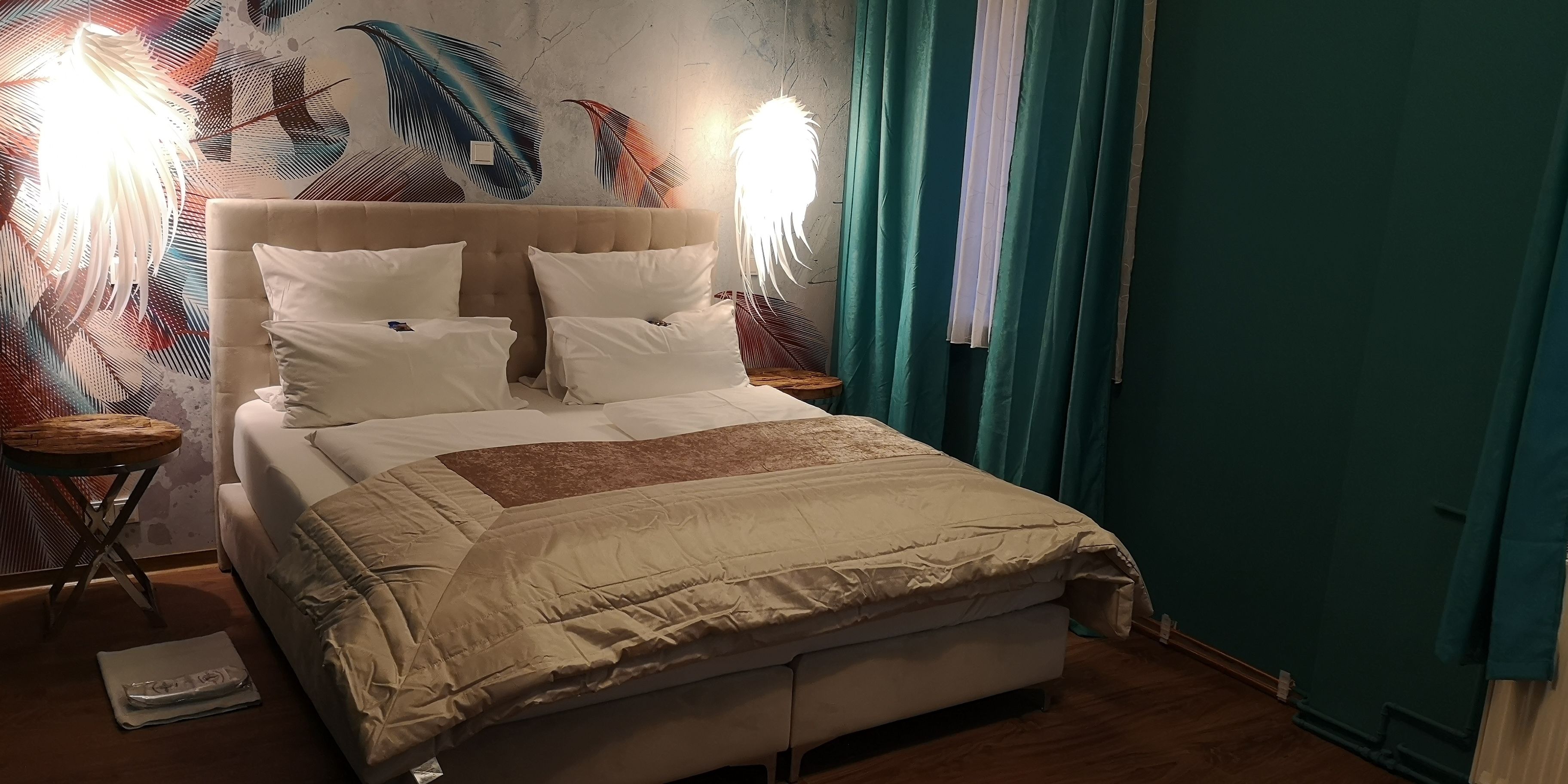 Una cama con ropa blanca y cabecera color beige, flanqueada por dos mesitas de noche con lámparas, frente a una pared con un mural de motivos vegetales.