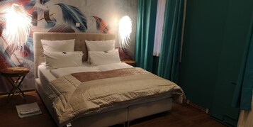 Una cama con ropa blanca y cabecera color beige, flanqueada por dos mesitas de noche con lámparas, frente a una pared con un mural de motivos vegetales.