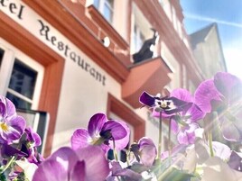 Um hotel com uma placa que diz 'Hotel Restaurant' e vista para flores roxas ao primeiro plano.