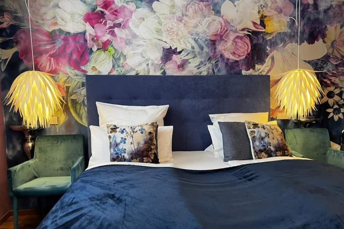 A bedroom with a large bed, two green armchairs, a floral wall mural, and two lamps with yellow shades.