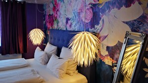 Quarto de hotel com cama, roupa de cama branca, um grande mural floral e duas luminárias pendentes.