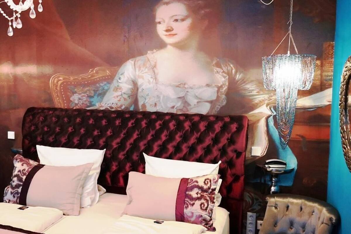 A bed with a patterned headboard, a chandelier, and a large portrait on the wall.