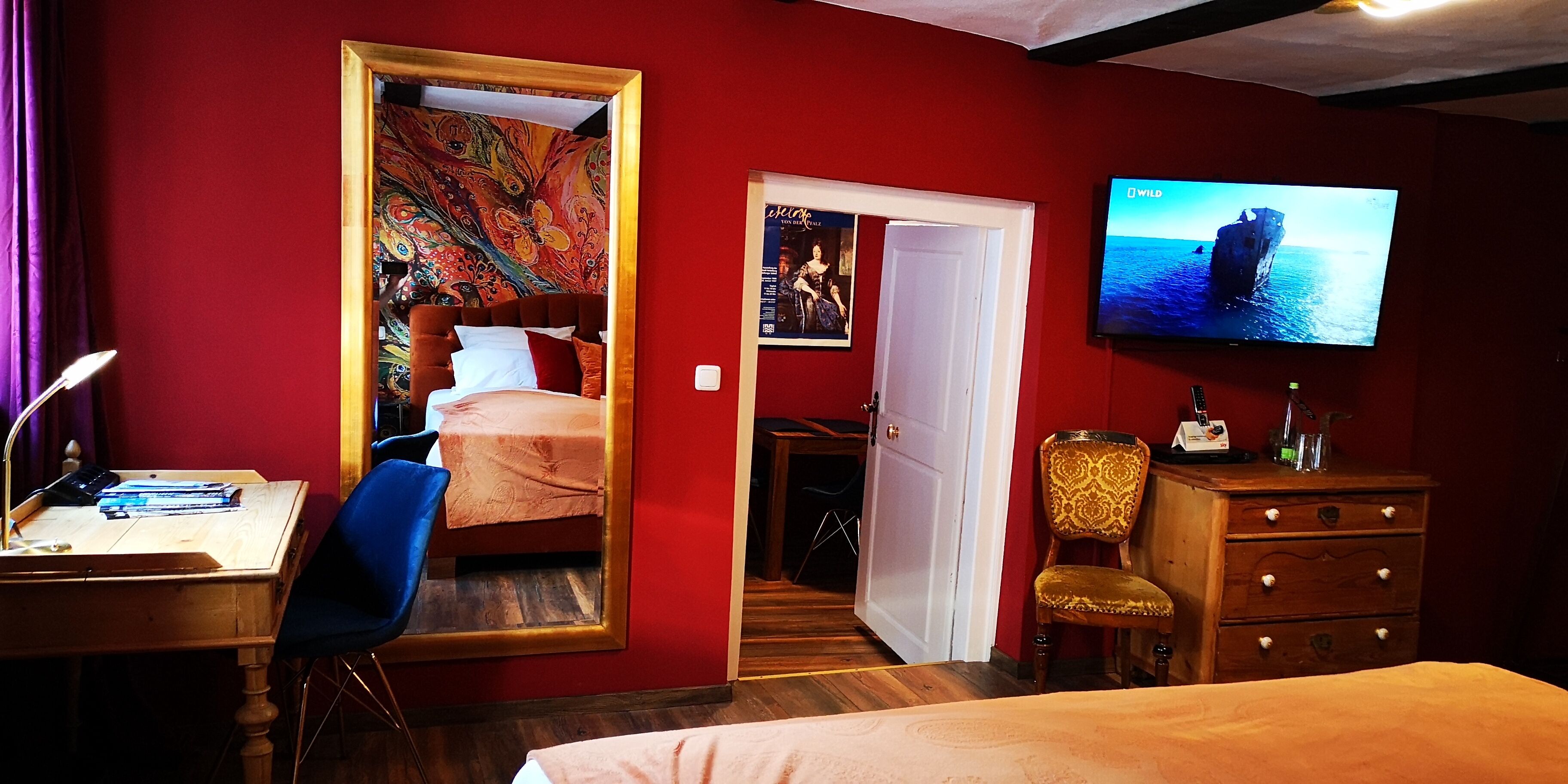 A hotel room with a bed, a desk with a chair, a television, a dresser, and a door leading to another room.