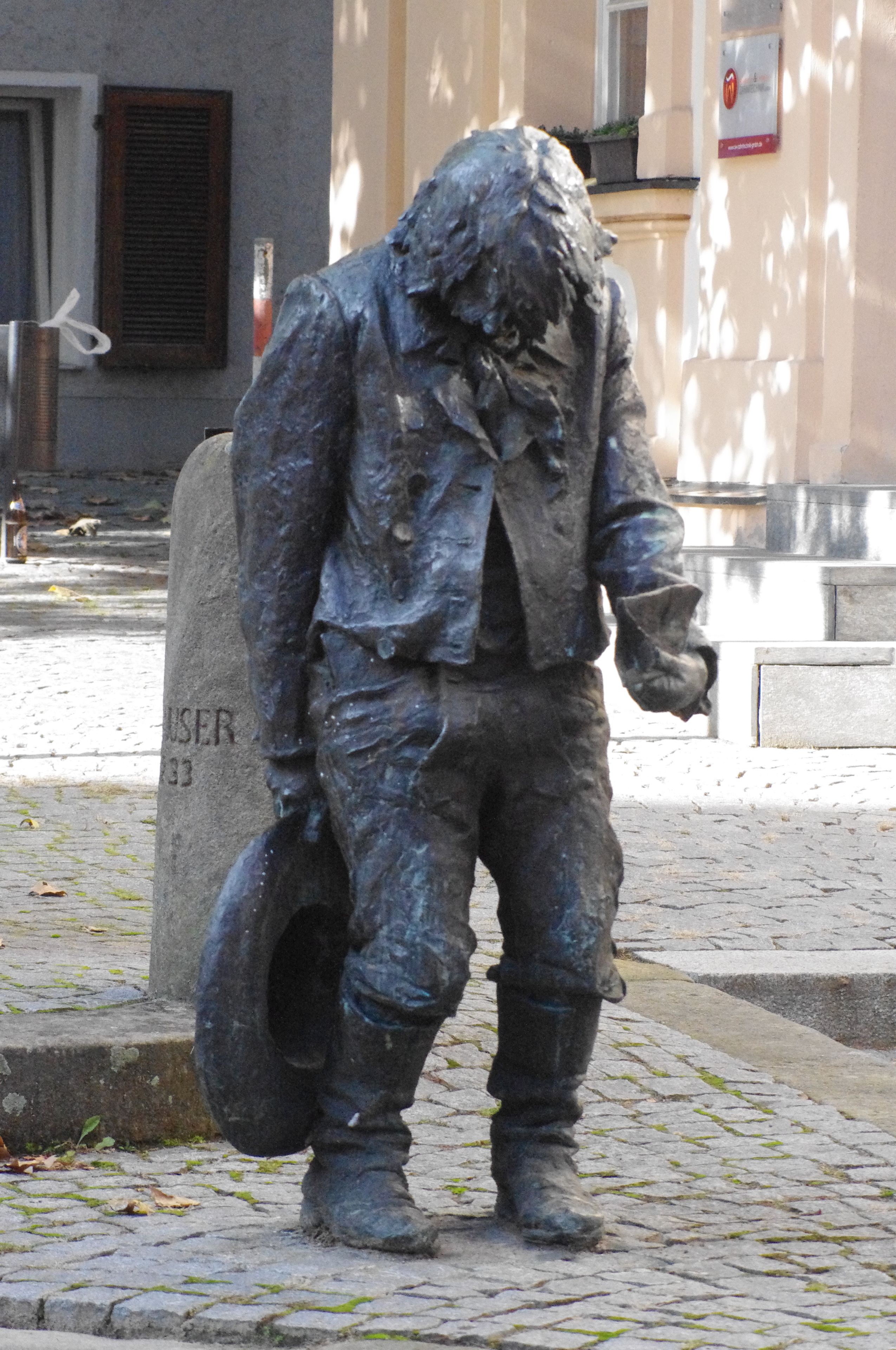 Uma estátua de uma pessoa vestida com traje histórico, segurando um capacete, em um pavimento de pedras irregulares, com um edifício ao fundo.