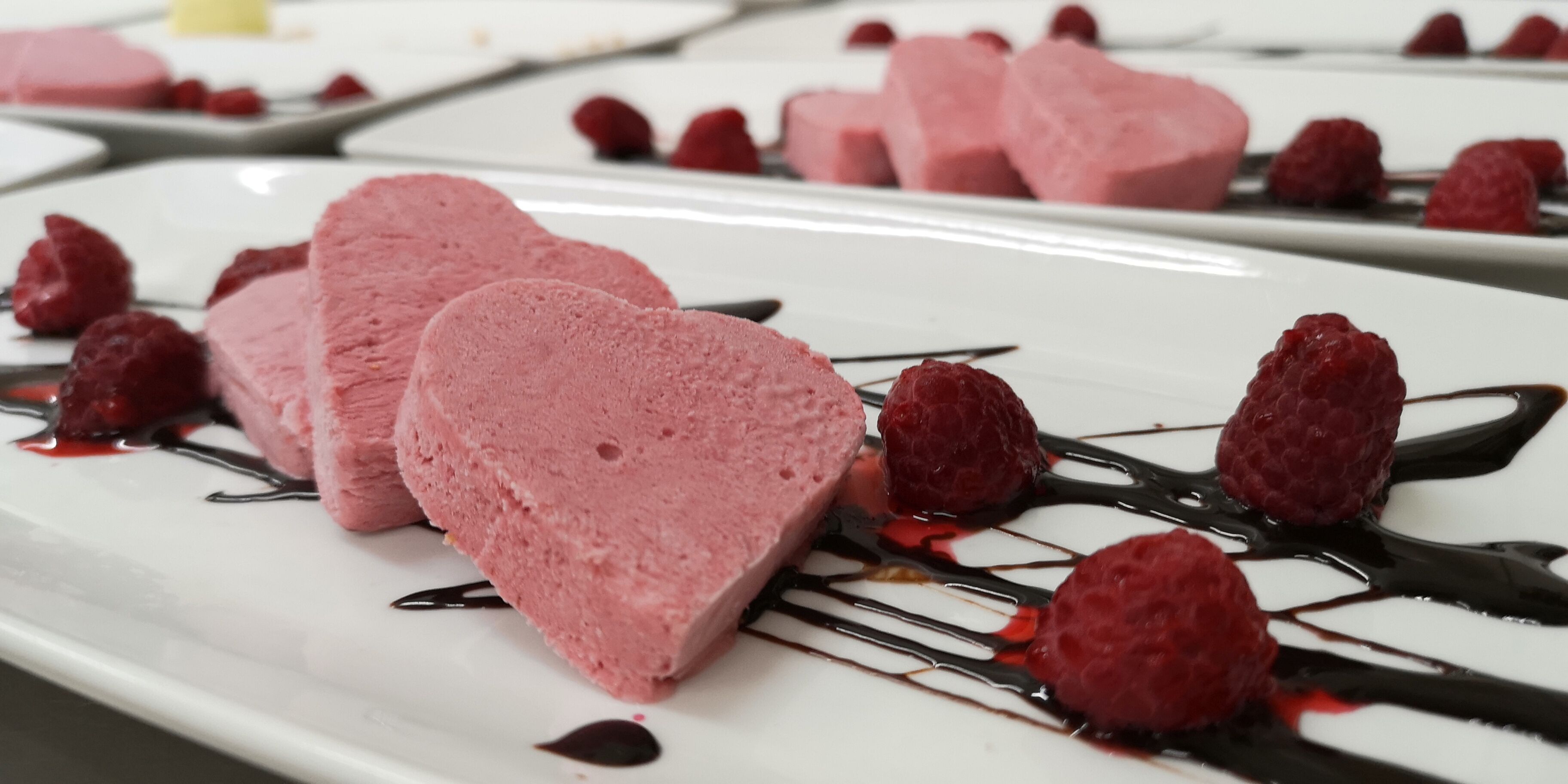 A dessert plate with heart-shaped pink ice cream and raspberries, garnished with chocolate sauce.