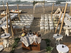 A beach wedding setup with white chairs, tables, and a wooden bench with a blanket.
