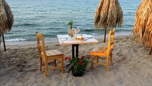 Een tafel aan het strand met twee houten stoelen, een tafelkleed en een potplant met rode bloemen.